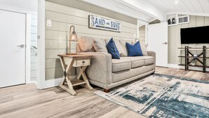 Un soggiorno con un divano beige, un tavolino di legno, un tappeto blu e bianco e un cartello con la scritta 'SAND AND SURF'.