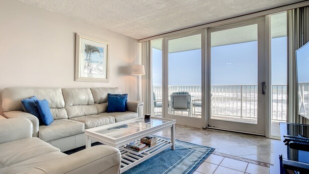 A living room with a beige sofa, a coffee table, and a view of the beach through large sliding glass doors. - Smyrna Beach Club 307 (New Smyrna Beach)