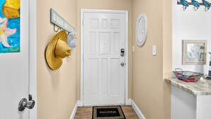 A hallway with a white door, a doormat, a wall-mounted hat rack, a mirror, and a shelf with decorative items. - NSB Sea Coast 401 (New Smyrna Beach)