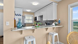 A kitchen with white cabinets, a marble countertop, and a view of the ocean through a window. - NSB Sea Coast 401 (New Smyrna Beach)