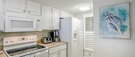 Cocina con armarios blancos, microondas, estufa, refrigerador y tostadora. Hay un cuadro en la pared.
