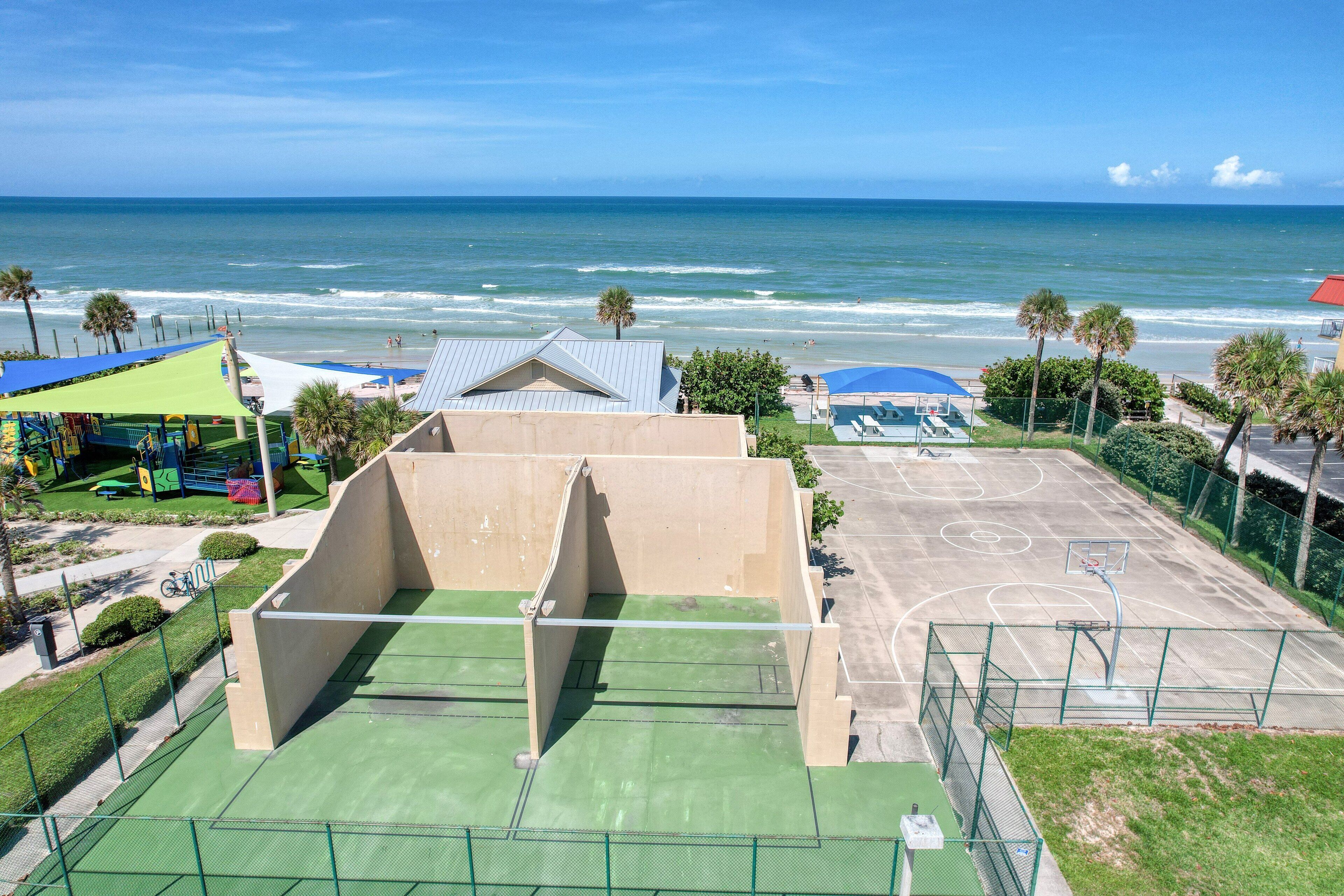 Ein Basketballplatz mit grüner Oberfläche neben einem Gebäude mit blauem Dach, mit Blick auf einen Strand mit Palmen und einem klaren Himmel.
