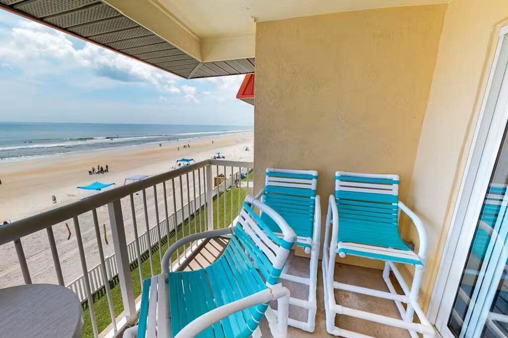 A balcony with two turquoise chairs and a table, overlooking a beach with people and a lifeguard tower.