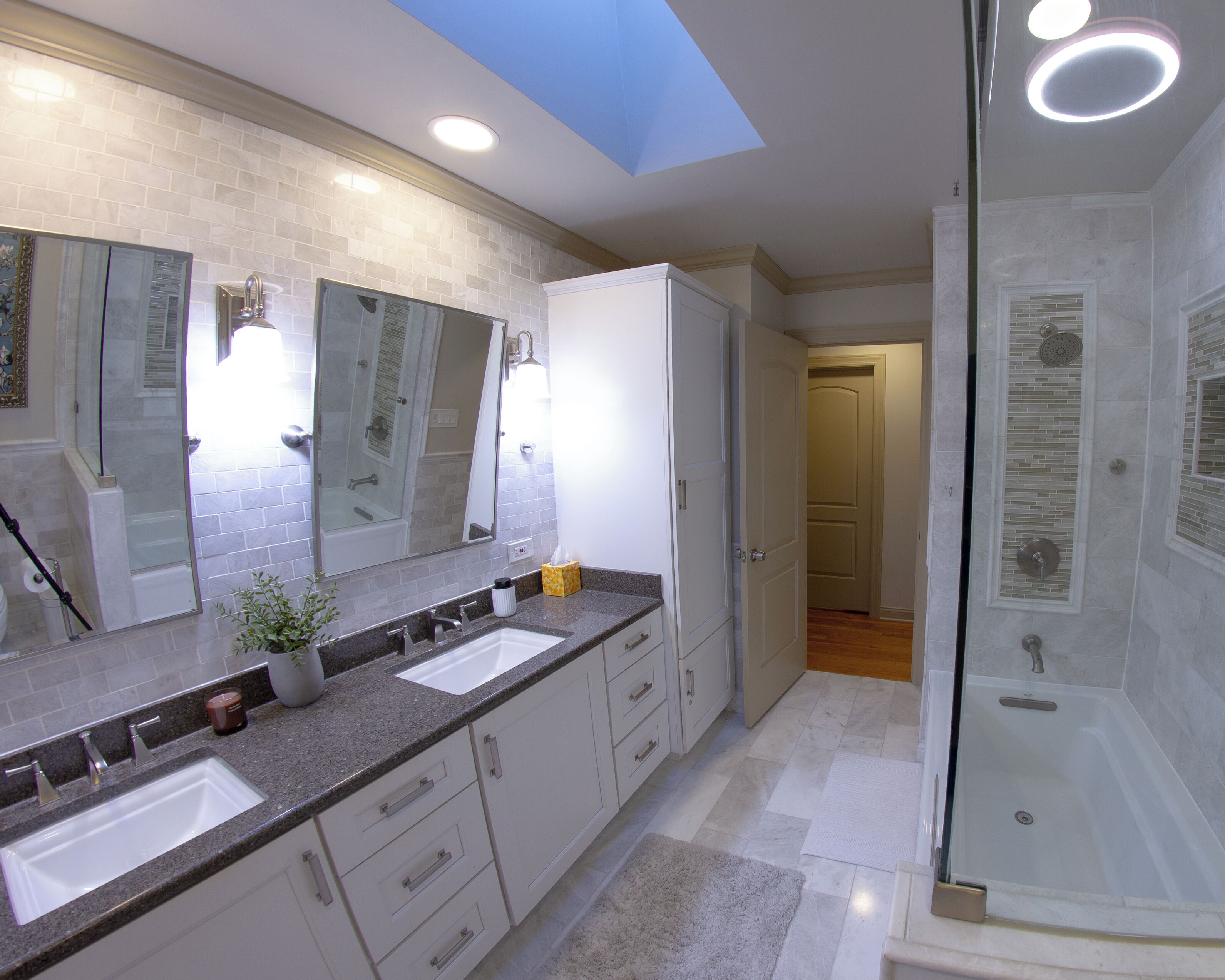 A modern bathroom with a large mirror, two sinks, a shower, and a skylight.
