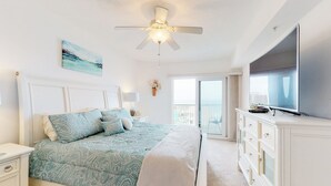 A bedroom with a bed, a nightstand, a dresser, a ceiling fan, a TV, and a window with a view. - Daytona Grande Towers - Ocean Paradise 702 (Daytona Beach Shores)