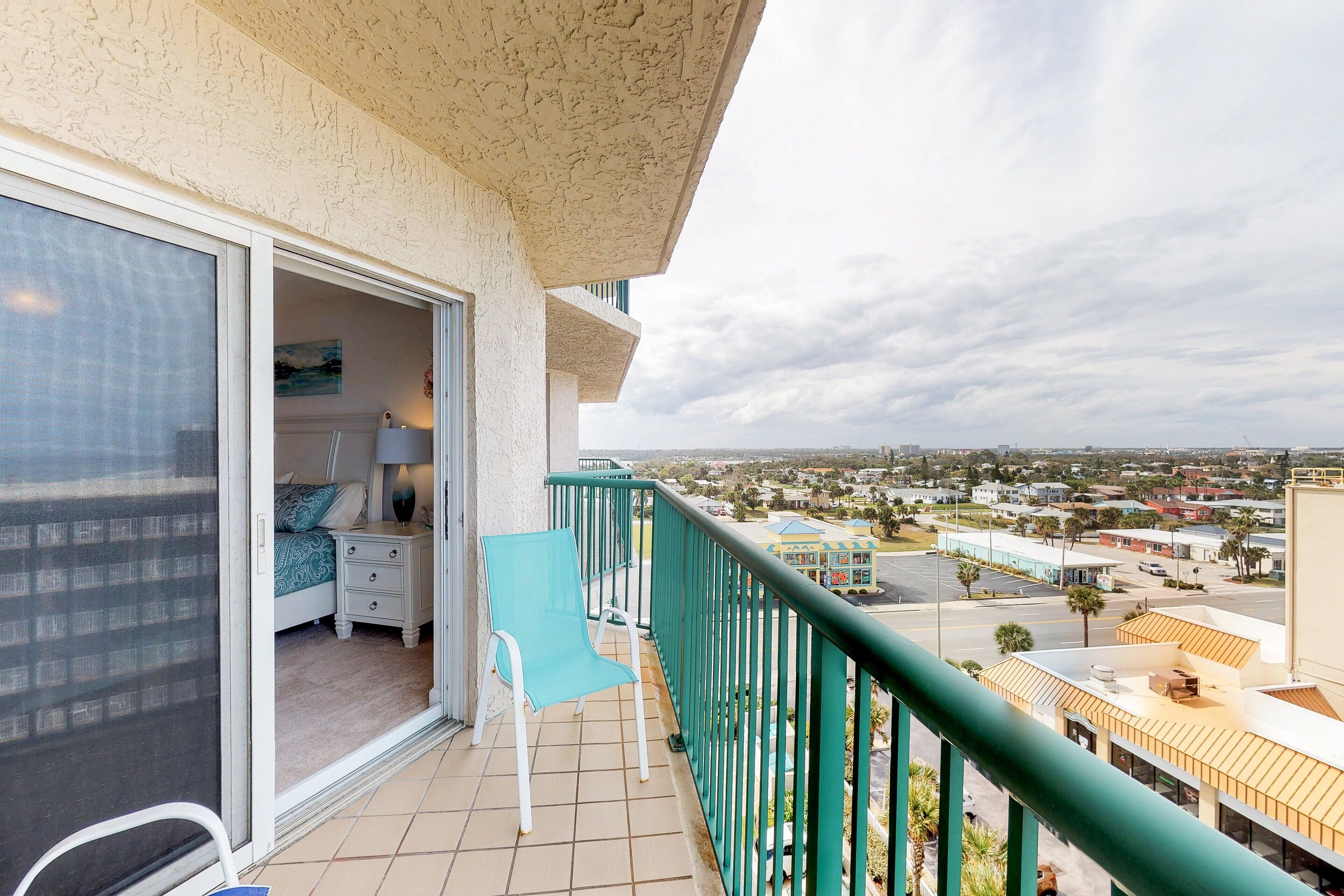 A balcony with a view of a cityscape, a bed with a blue and white patterned bedspread, a small table with a lamp, and a blue chair.
