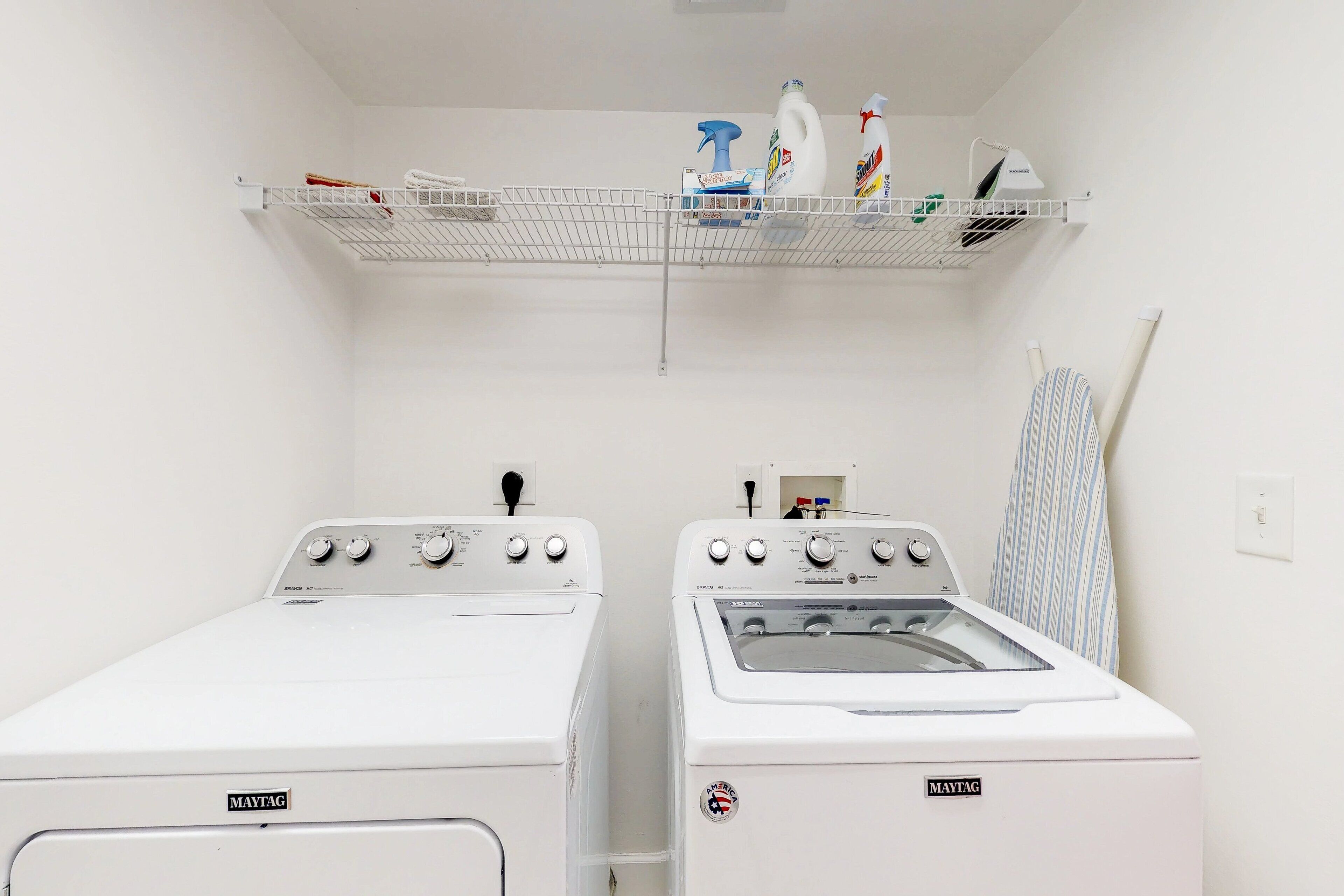 A laundry room with two Maytag washing machines, a shelf with cleaning supplies, and an ironing board.