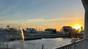 Un balcon offrant une vue sur un coucher de soleil se reflétant sur une étendue d’eau, une balustrade et un verre de vin rouge.