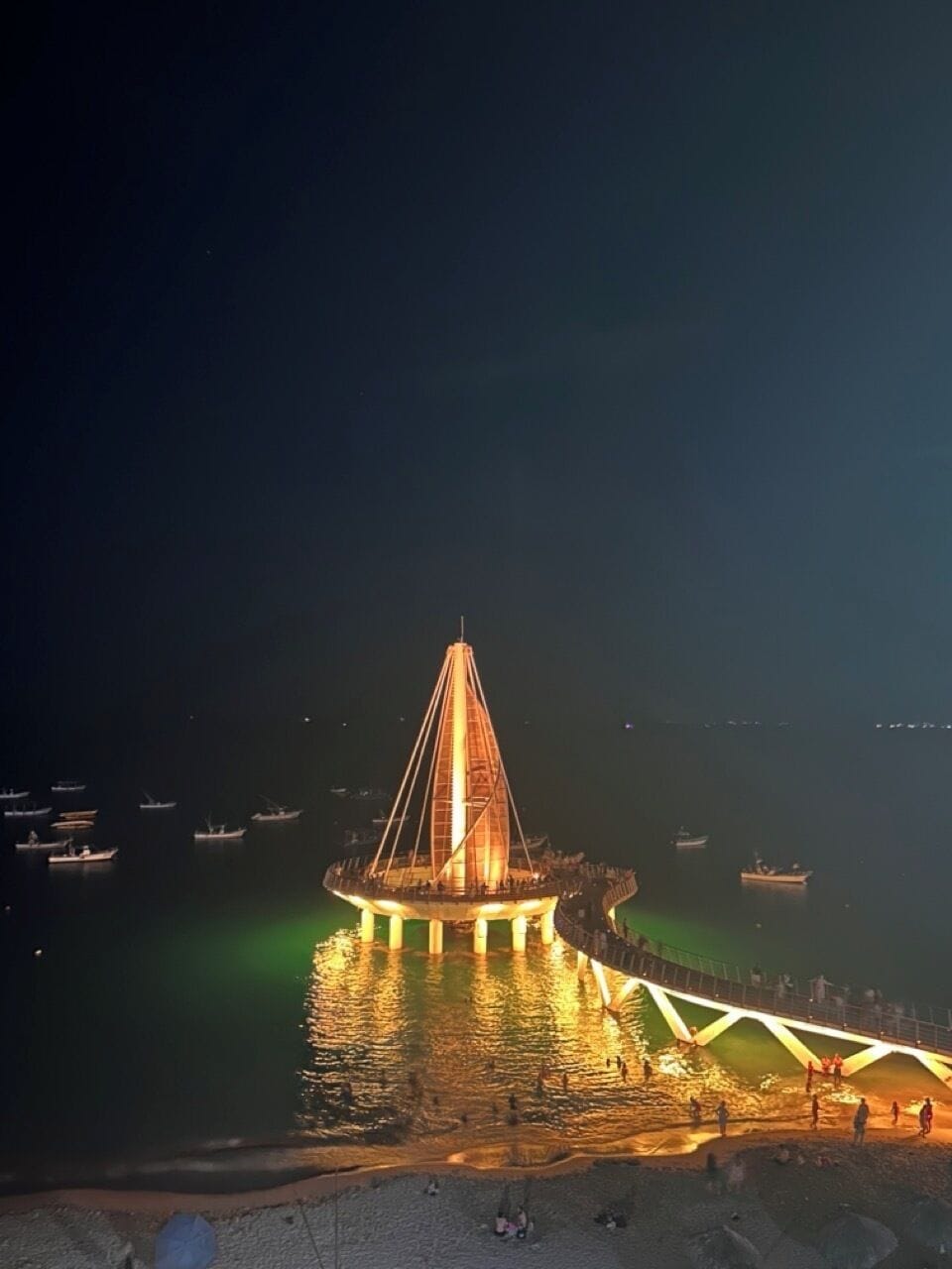 Vista nocturna de un puente con una estructura triangular, rodeado de aguas tranquilas y barcos.