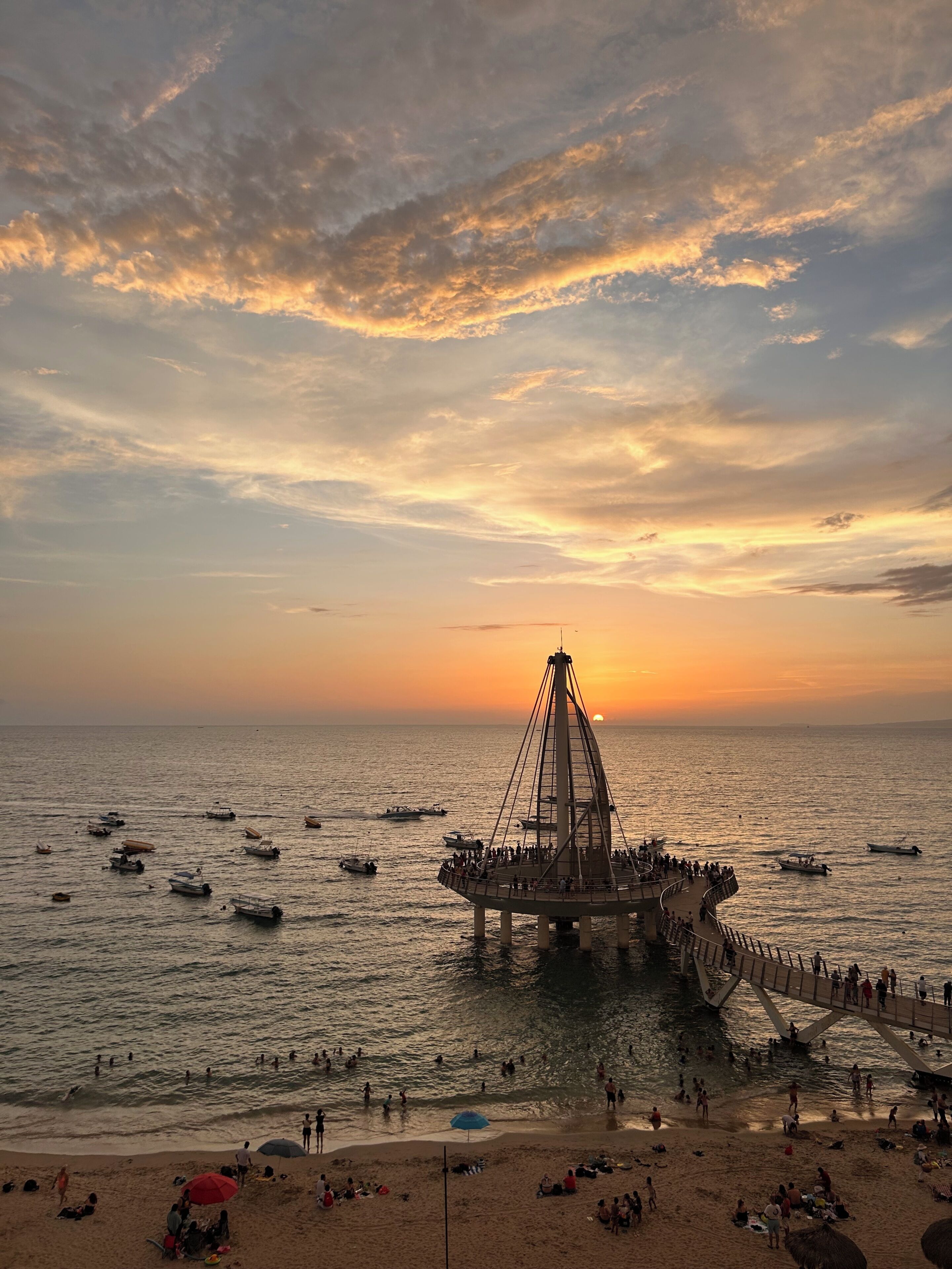 Eine Küstenansicht mit einer Brückenstruktur über dem Wasser, Strandbesuchern und Booten.