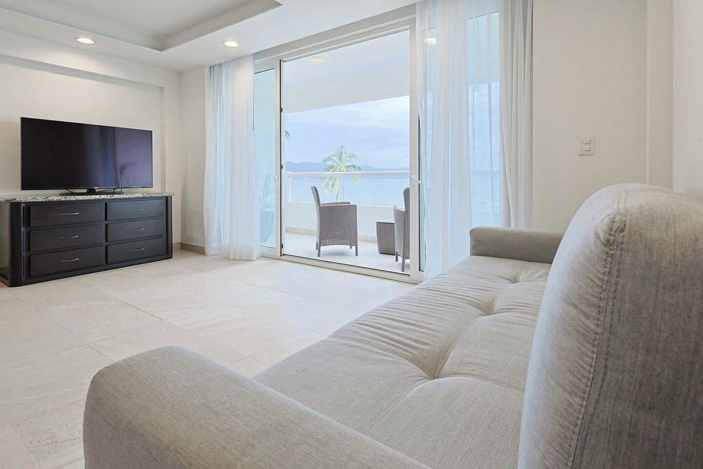 A modern living room with a sofa, a flat-screen TV, and a sliding glass door leading to a balcony with outdoor furniture.