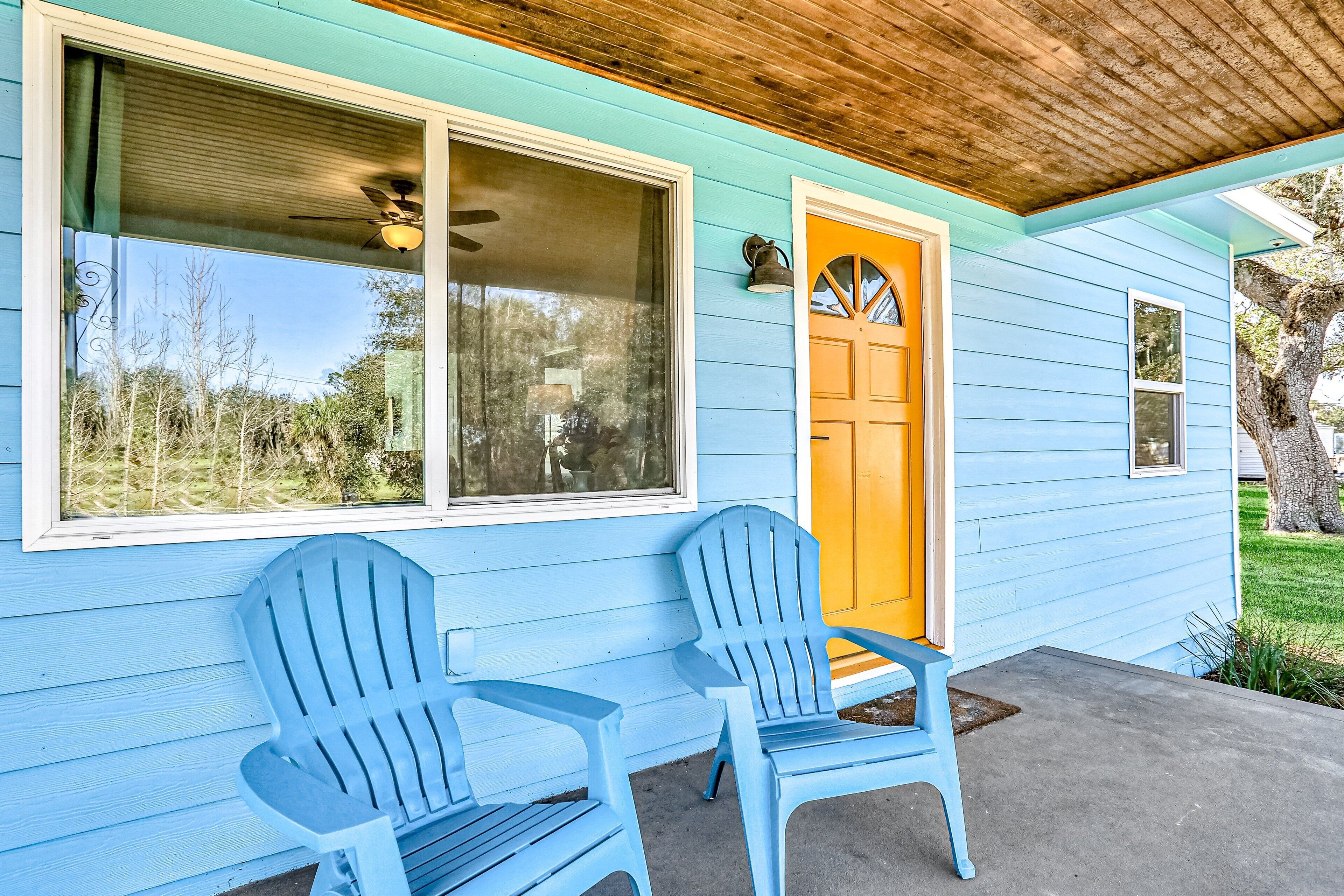 A light blue house with a yellow door, two blue Adirondack chairs, and a ceiling fan.