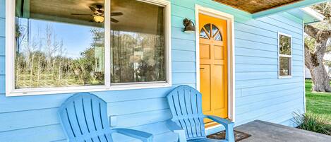 A light blue house with a yellow door, two blue Adirondack chairs, and a ceiling fan.