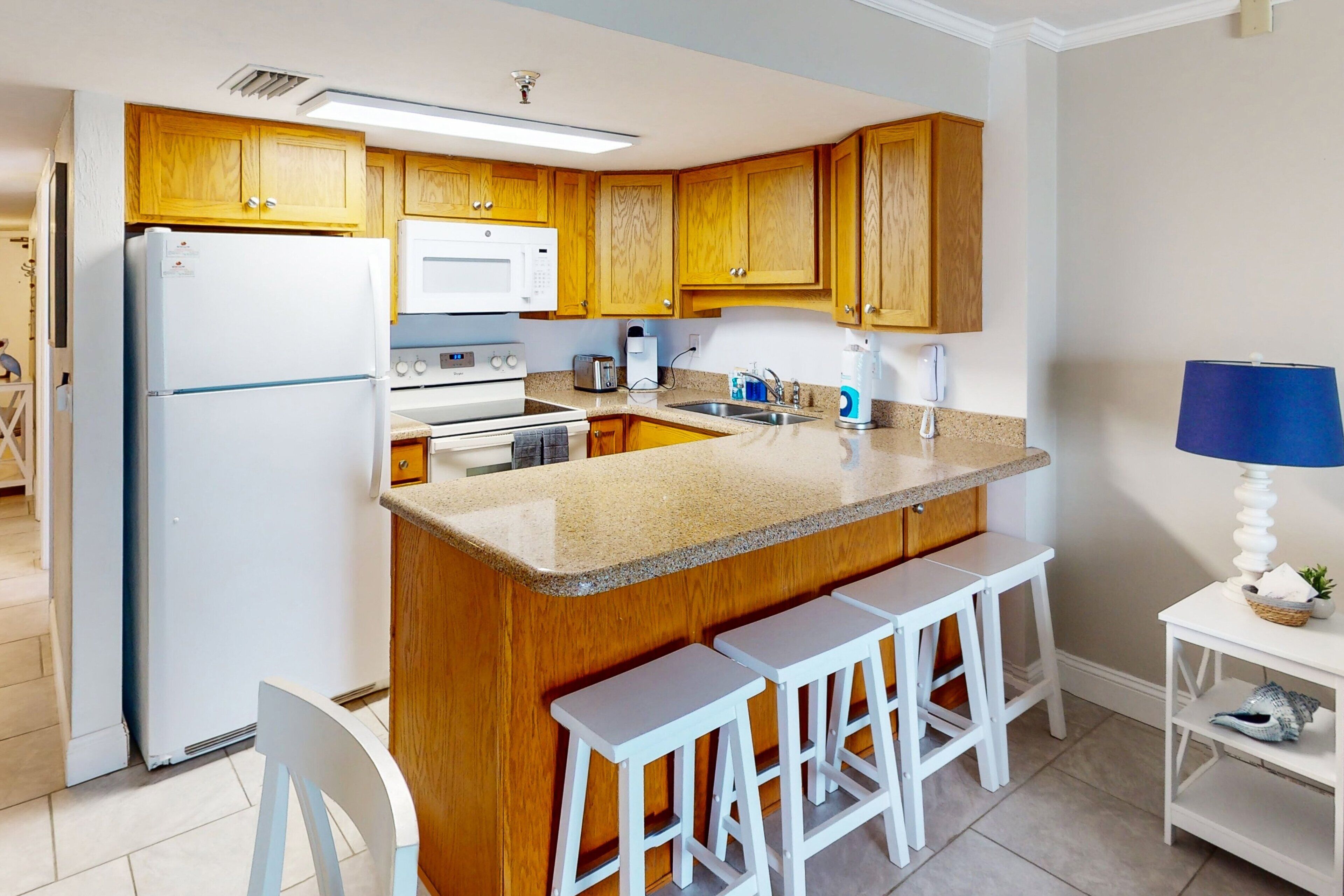 A modern kitchen with wooden cabinets, a white refrigerator, a microwave, and a granite countertop. There are three white bar stools and a small white table with a lamp.