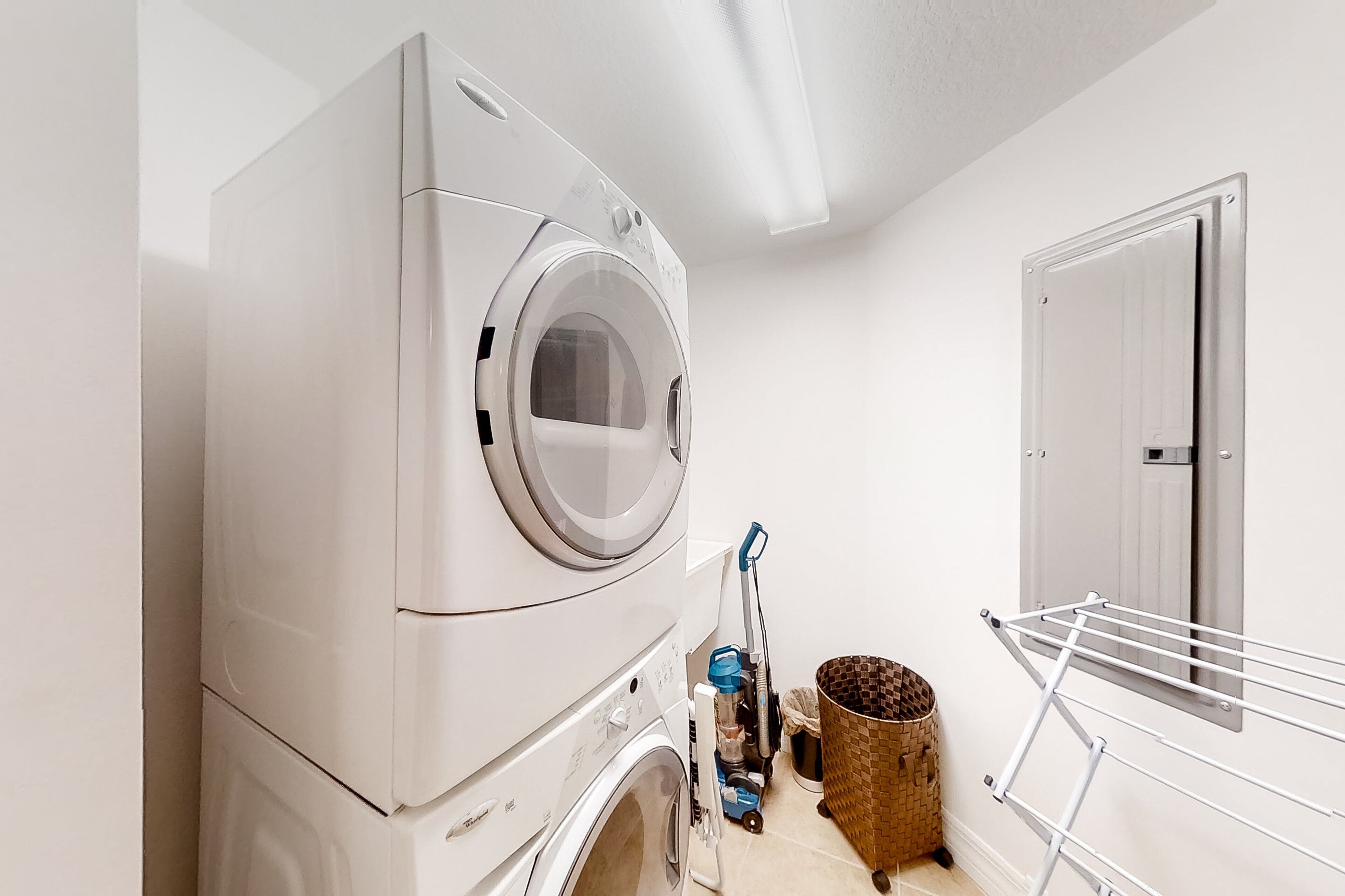 A compact laundry room with a front-loading washing machine and dryer, a drying rack, a mop, and a trash bin.