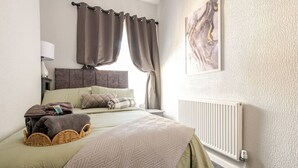 Un dormitorio con cama, almohadas, mesita de noche, lámpara, radiador y ventana con cortinas.