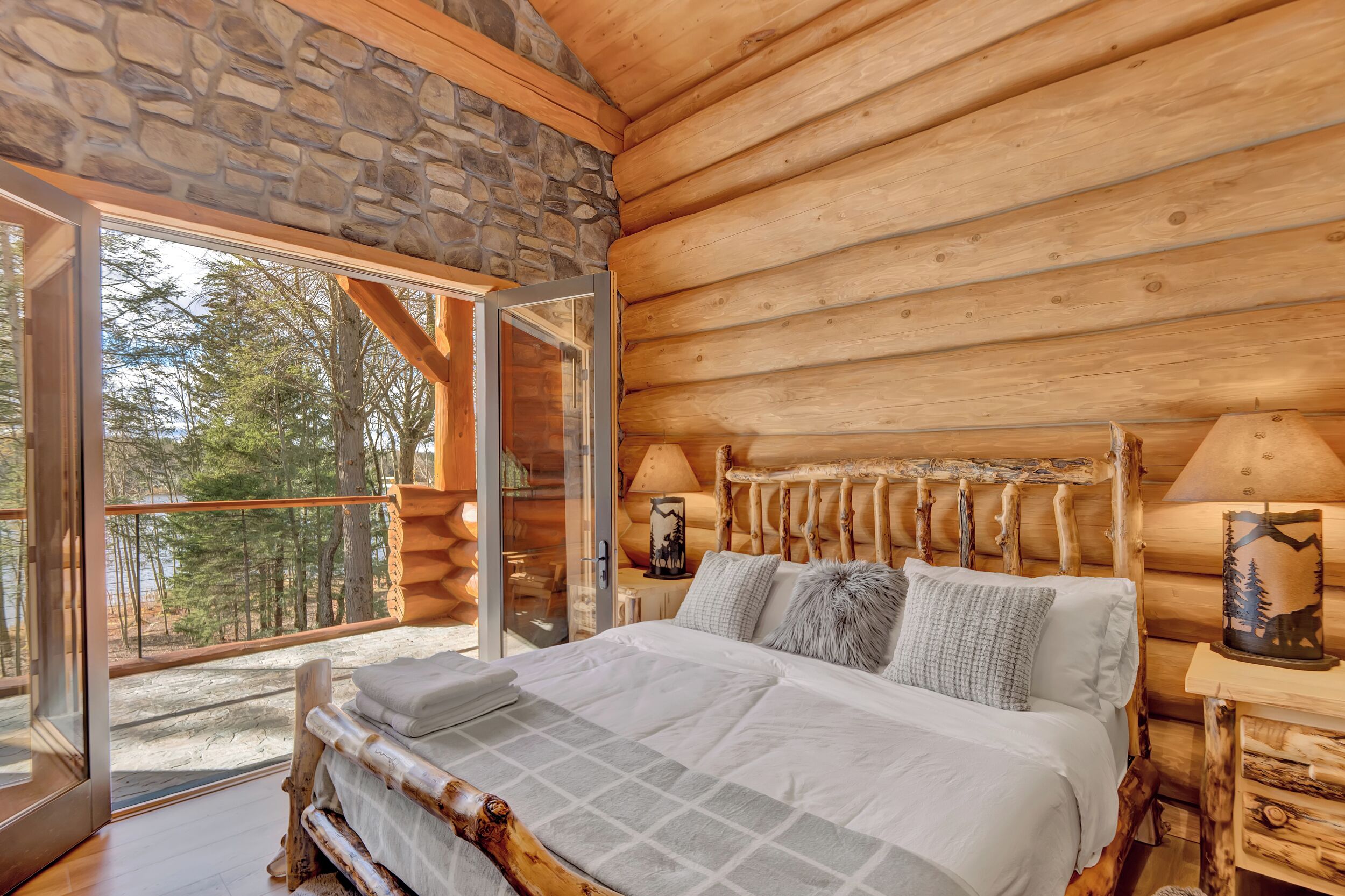A log cabin bedroom with a stone wall, wooden ceiling, and a large bed with pillows and a lamp.