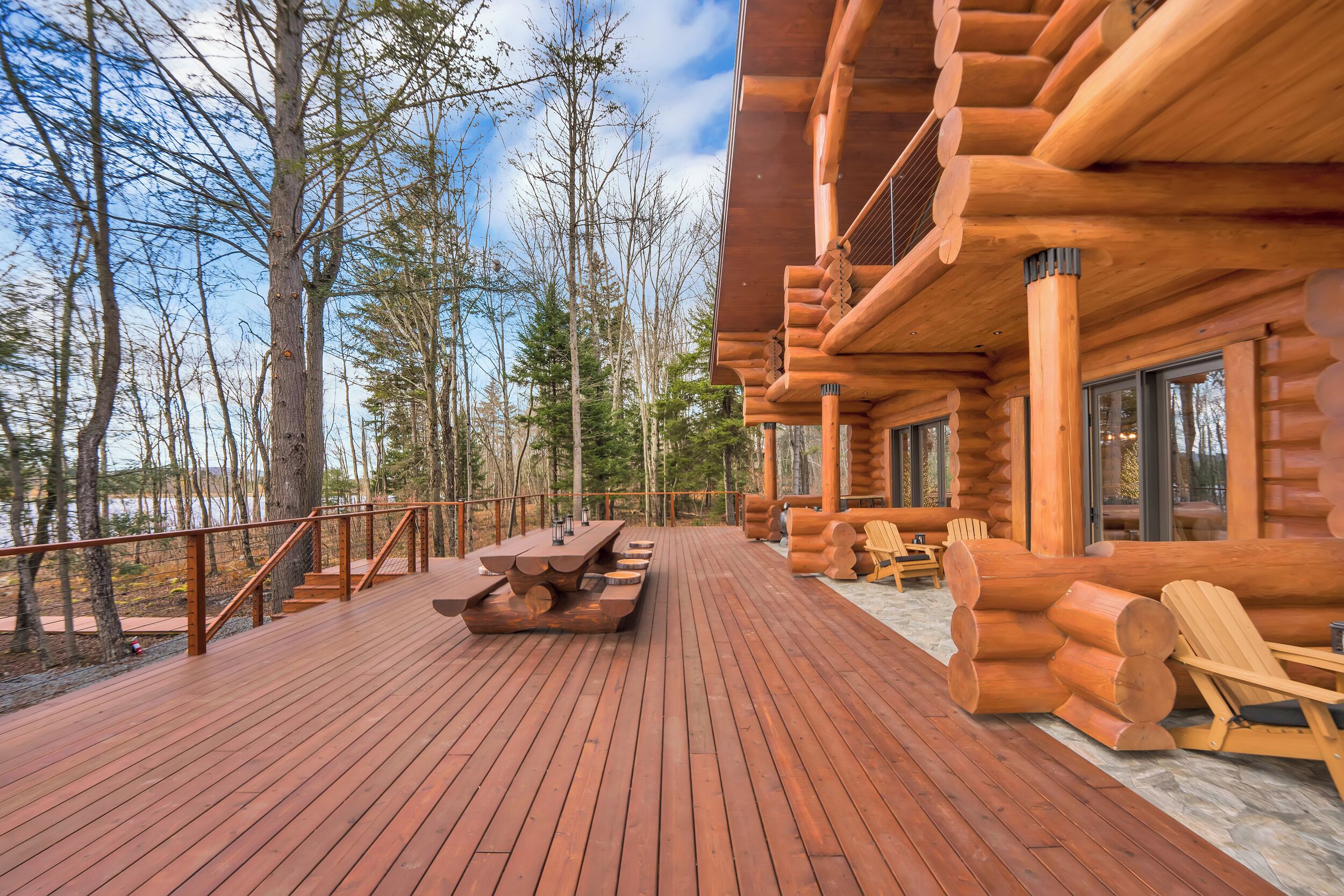 A wooden deck with a picnic table and chairs, surrounded by trees and a lake.