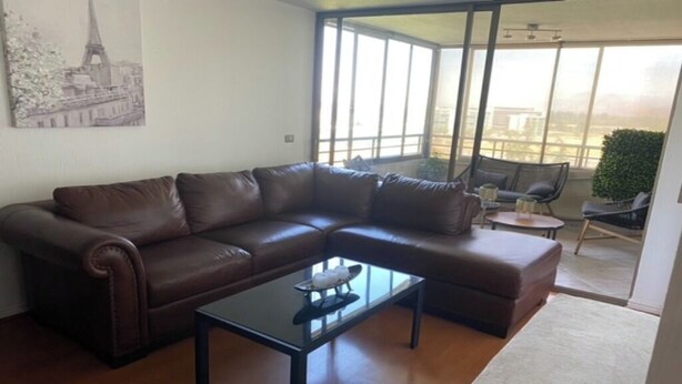 Ein Wohnzimmer mit braunem Ledersofa, einem Glastisch und Blick auf einen Balkon durch große Fenster.