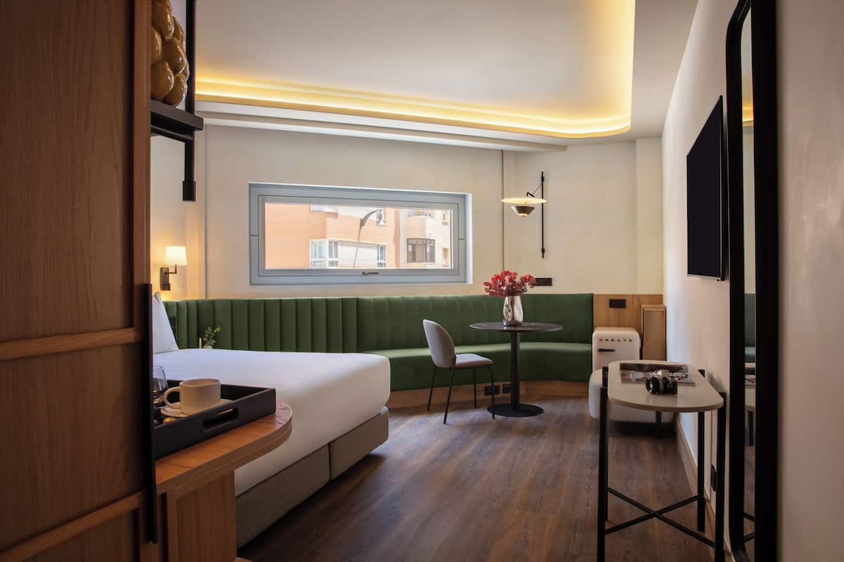 A hotel room with a bed, a green sofa, a small table with a vase, and a window with a view of buildings.