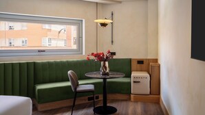 Et moderne værelse med en grøn sofa, et lille rundt bord med en blomstervase, en kompakt hvid enhed og et vindue med udsigt til bygninger.