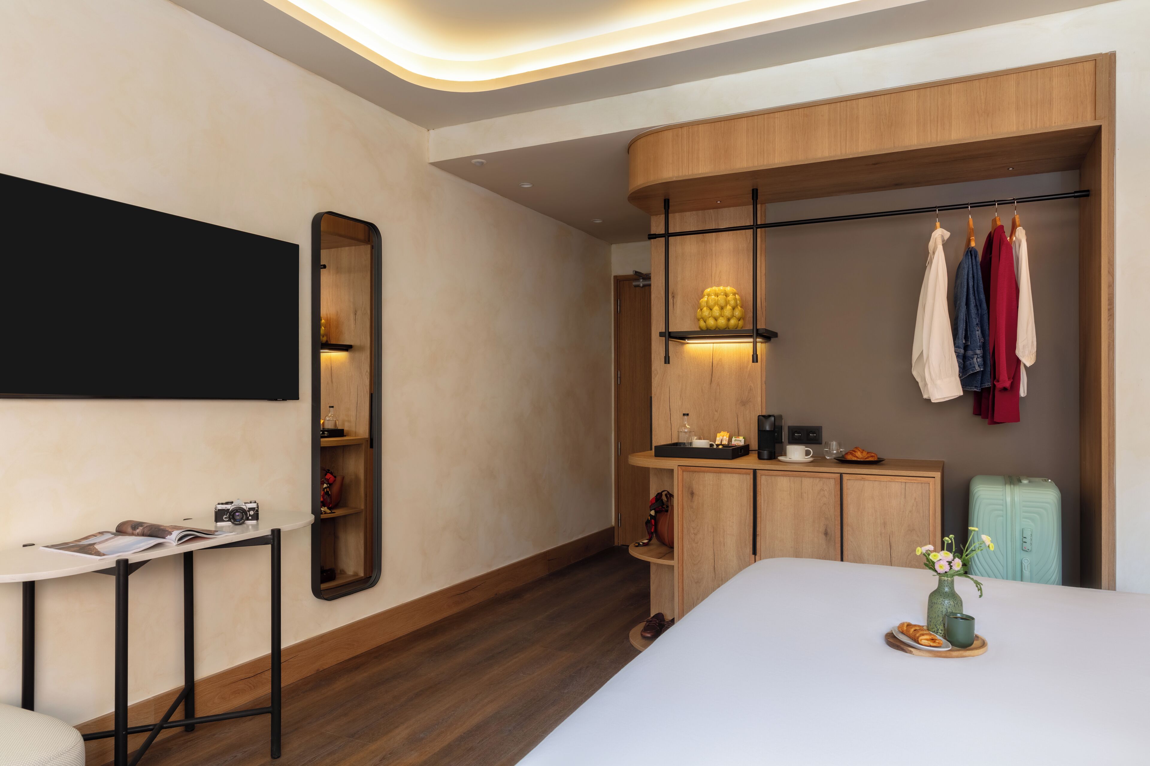 A modern hotel room with a large bed, a bedside table with a camera, a mirror, a wardrobe with clothes hanging, and a small table with a vase and a plate.