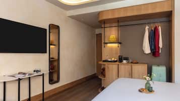 A modern hotel room with a large bed, a bedside table with a camera, a mirror, a wardrobe with clothes hanging, and a small table with a vase and a plate.