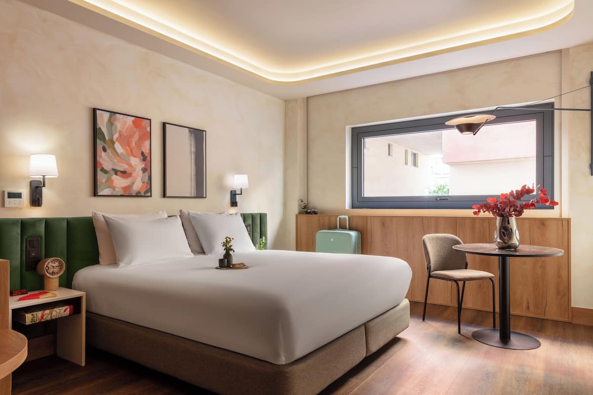 A modern hotel room with a large bed, bedside tables, a small table with a vase, and a window with a view.
