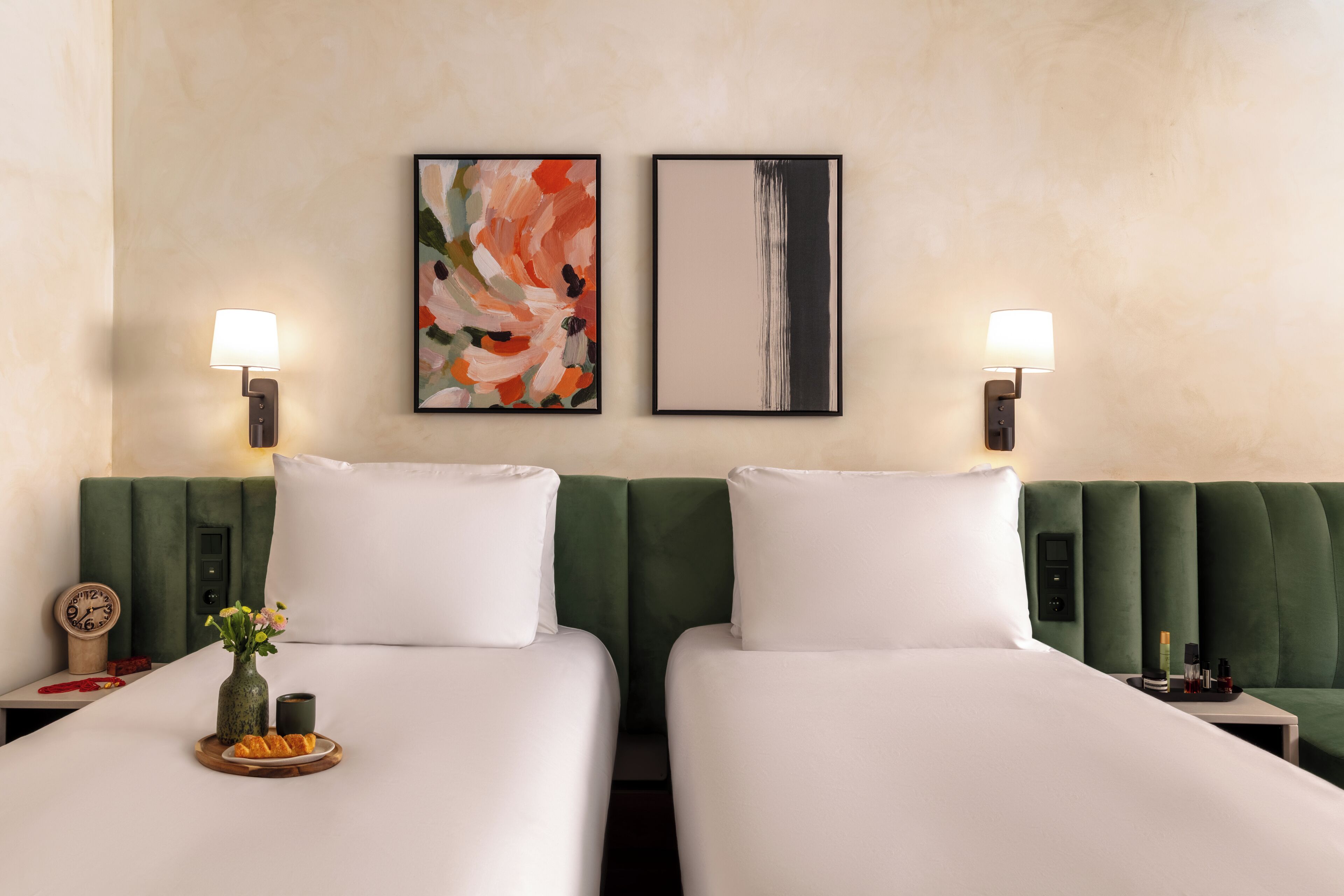 A hotel room with two beds, green headboards, white bedding, two wall-mounted lamps, and a painting on the wall.