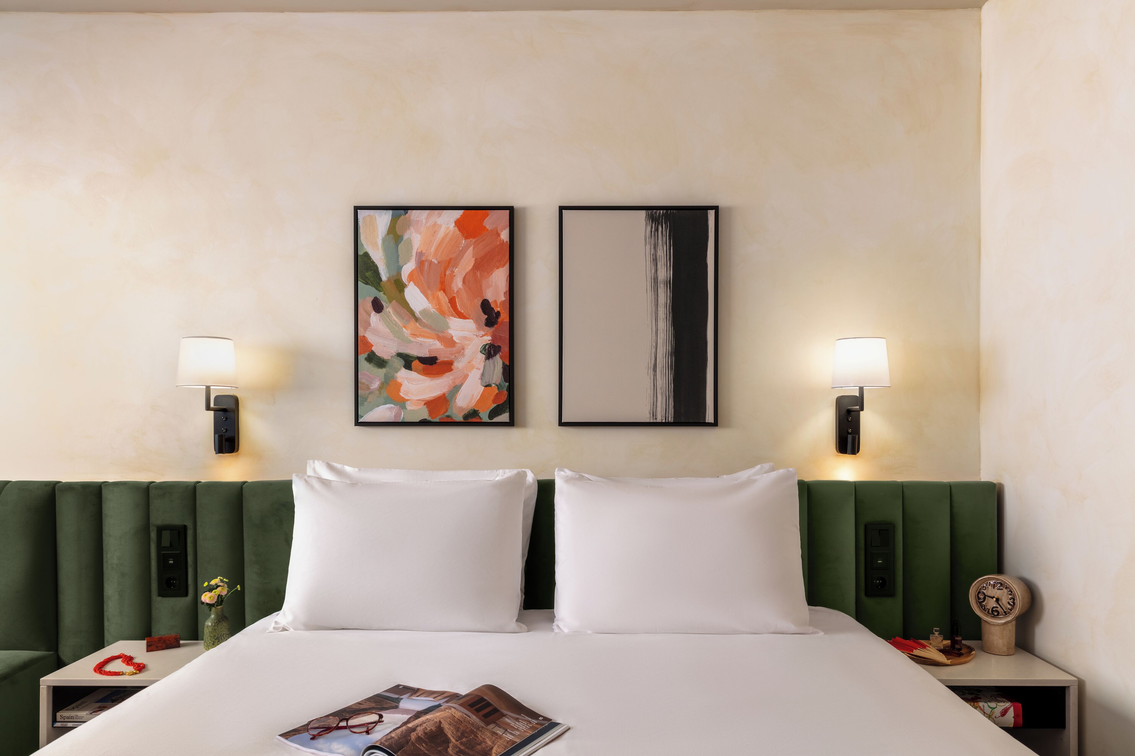 A hotel room with a green upholstered headboard, white bedding, two framed artworks, bedside lamps, a clock, and a small vase with flowers.
