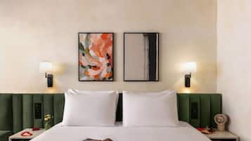 A hotel room with a green upholstered headboard, white bedding, two framed artworks, bedside lamps, a clock, and a small vase with flowers.