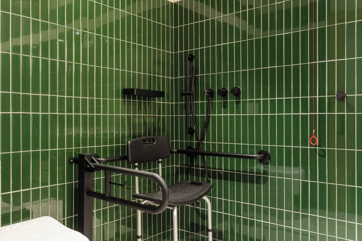 A bathroom with green tiled walls, a black shower chair, and a black towel rack.