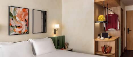 Habitación de hotel con cama, mesita de noche, lámpara, un reloj de pared, repisa con una prenda roja y una revista sobre la cama.