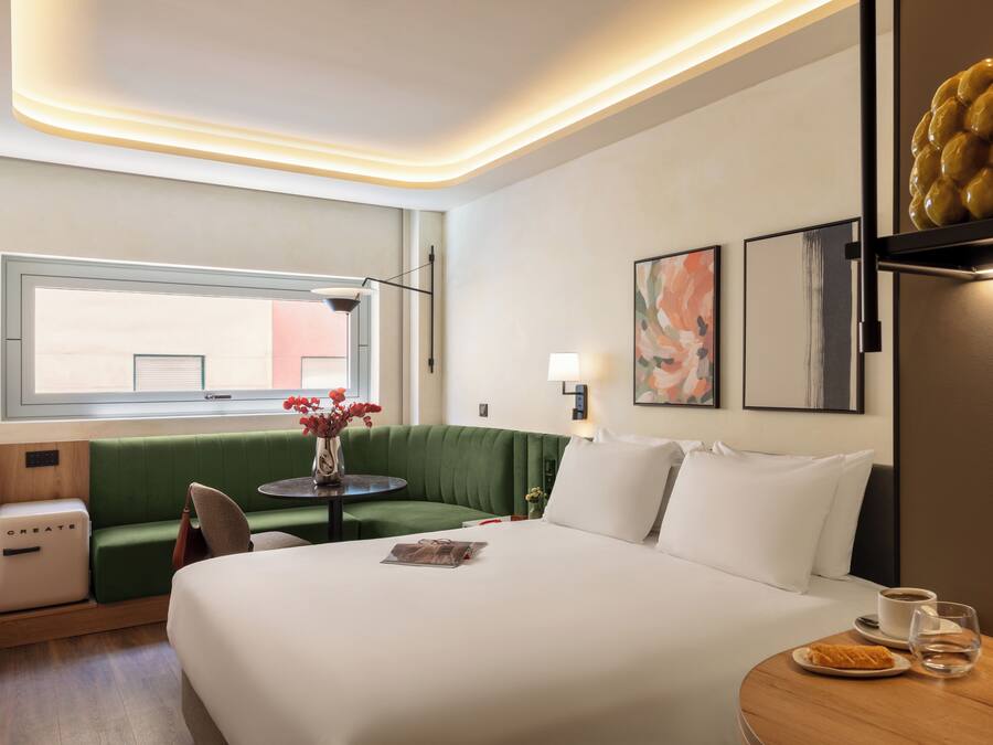 Habitación de hotel con sofá verde, cama, una mesita de noche con lámpara y ventana con vistas.