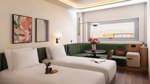 Et moderne hotelværelse med to senge, en grøn sofa, et lille bord med en vase og et vindue med udsigt.