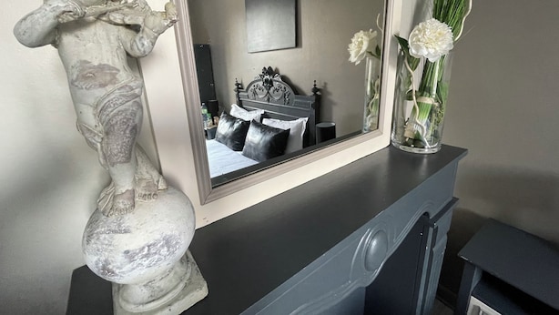 Una statua in marmo su un comò, uno specchio che riflette una camera da letto con un letto e un comò, e un vaso con fiori bianchi sul comò.