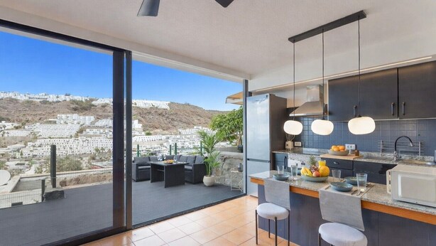 A modern kitchen with a large island, bar stools, and a view of a hillside town. - Arizona's Bay Holiday Home with Sea Views, Hot Tub, and Private Terrace (Mogán)