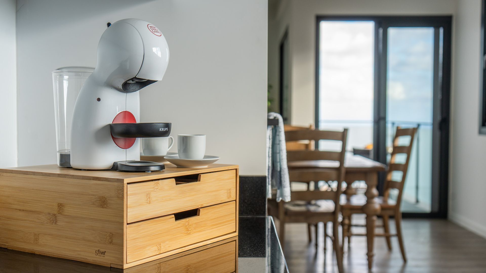 A modern kitchen with a coffee machine on a wooden cabinet, a glass container, and a cup.