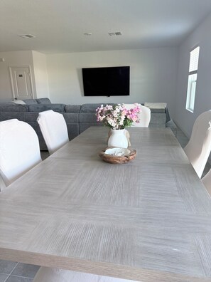 Ein moderner Essbereich mit einem grau strukturierten Tisch, weißen Stühlen, einer weißen Vase mit rosa Blumen und einem grauen Sofa im Hintergrund.