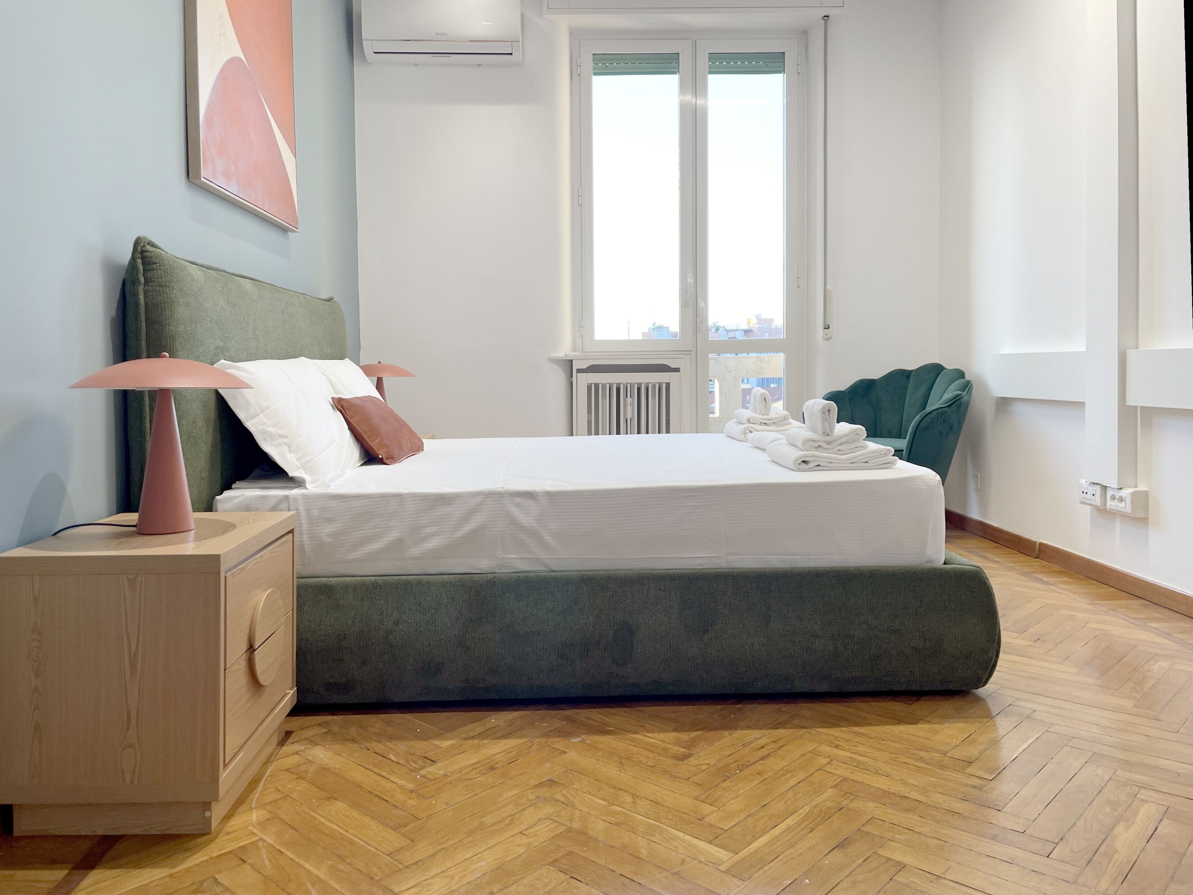 A modern hotel room with a wooden herringbone floor, a green upholstered bed, a bedside lamp, a radiator, and a view through the window.