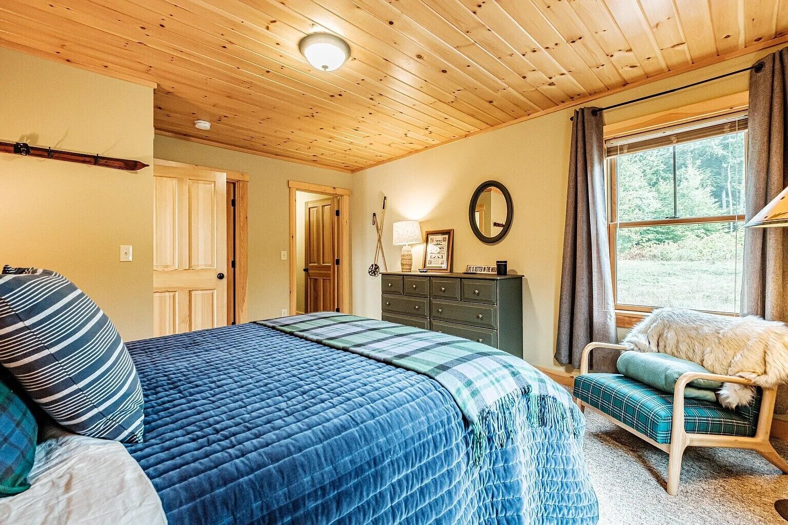 A cozy bedroom with a large bed, a dresser, a chair, and a window with a view of trees.