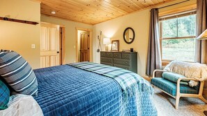 Ein gemütliches Schlafzimmer mit einem großen Bett, einer Kommode, einem Sessel und einem Fenster mit Blick auf Bäume.