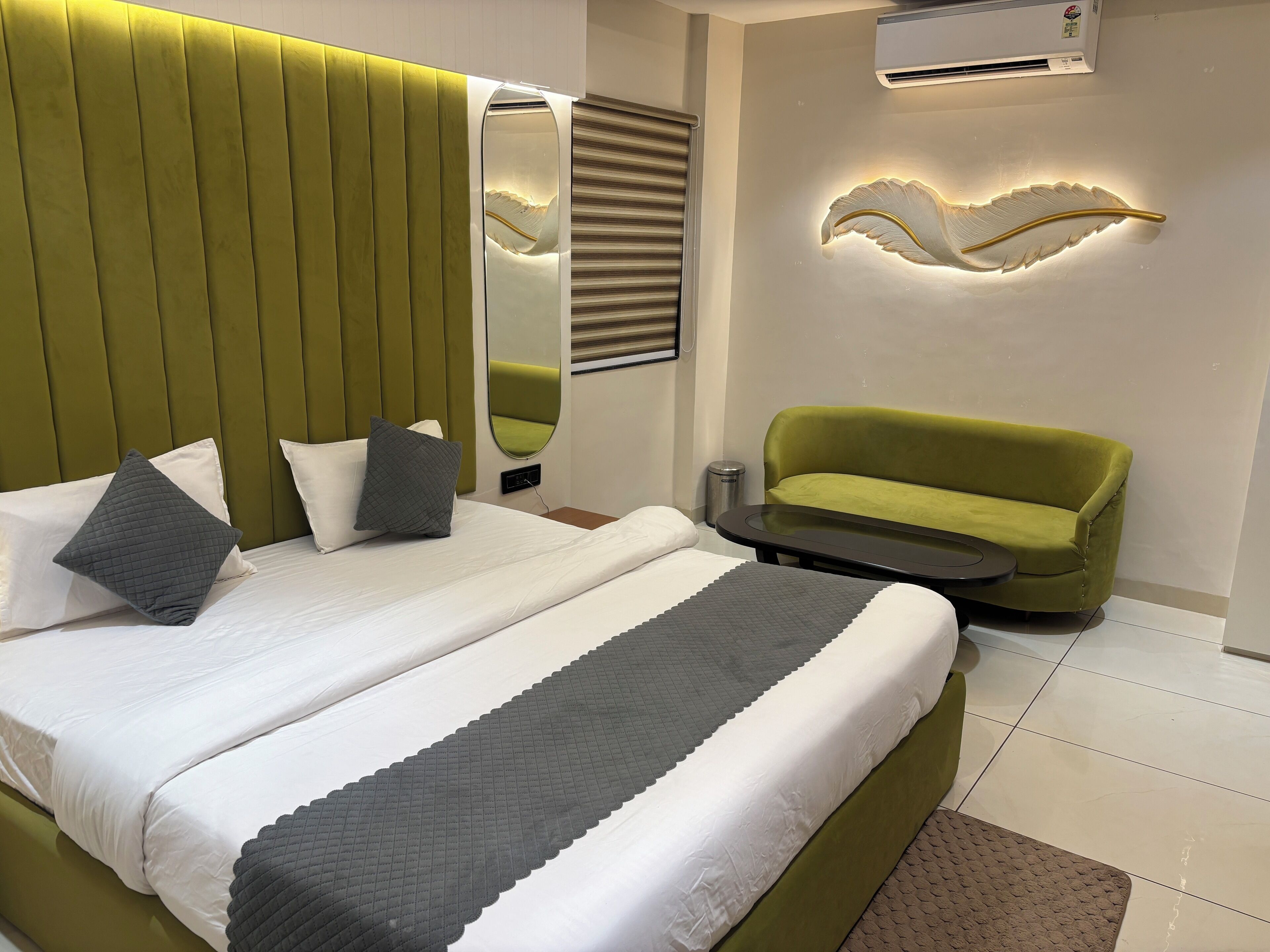 A modern hotel room with a large bed, a green sofa, a mirror, and an air conditioning unit.