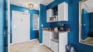 A compact kitchen with blue walls, white cabinets, a microwave, a coffee maker, and a trash bin. - Oceanfront Inn Room #21- Ocean View Balcony, King Bed (Fort Pierce)