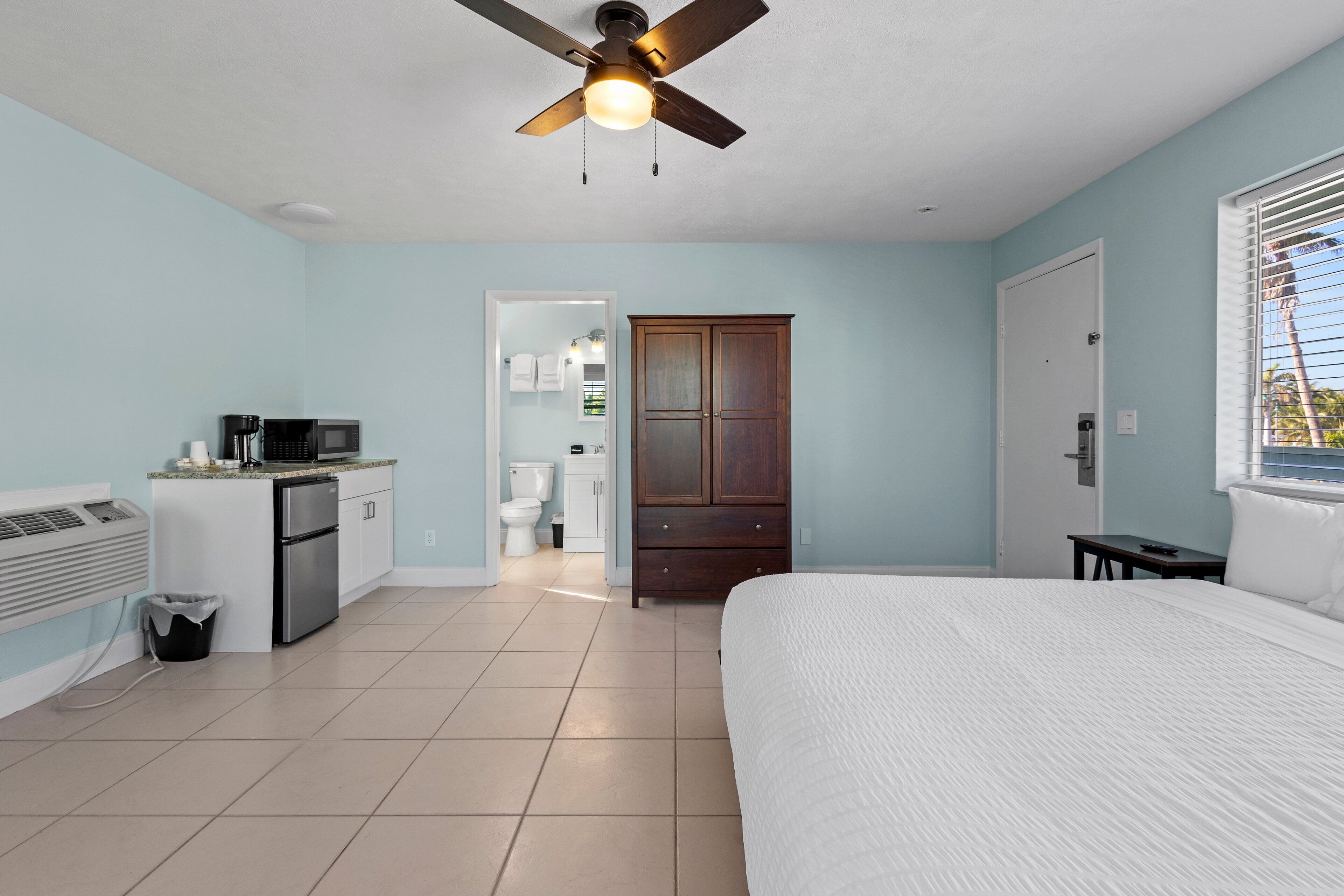 Une chambre à coucher avec un lit, un ventilateur de plafond, un coin cuisine et une salle de bain visible à travers une porte ouverte.