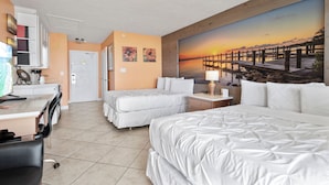 Une chambre d’hôtel avec deux lits, un bureau, une chaise et un tableau représentant un coucher de soleil sur une jetée.