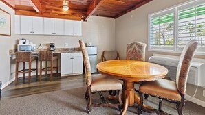 A dining area with a round wooden table and four chairs, a kitchen with white cabinets and a refrigerator, and a window with blinds. - Oceanfront Inn Suite #6- King Bed (Fort Pierce)