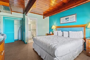 A bedroom with a large bed, a wooden ceiling, a blue wall, a wooden nightstand, and a door leading to a bathroom. - Oceanfront Inn Room #7 - Partial Ocean View - King Bed (Fort Pierce)