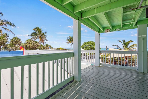 A covered wooden deck with white railings, palm trees, and a clear blue sky. - Oceanfront Inn Room #7 - Partial Ocean View - King Bed (Fort Pierce)