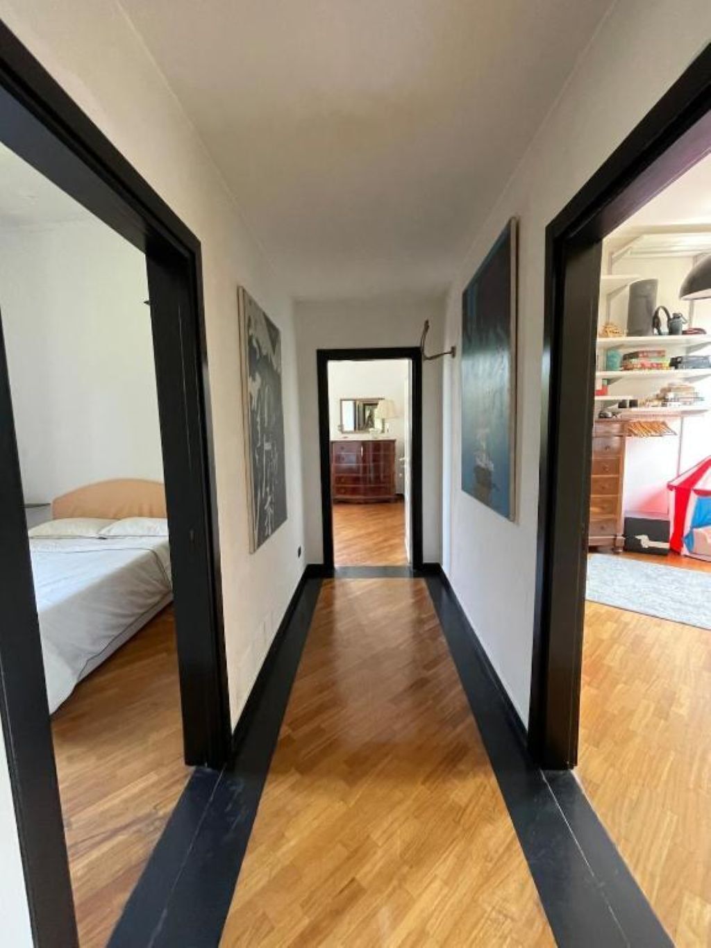 Een gang met houten vloeren en zwarte accenten, die leidt naar een slaapkamer met een bed en een badkamer met toilet en wastafel.